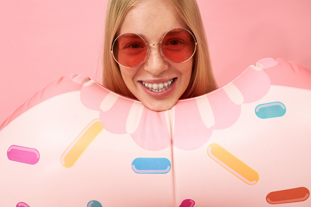 Taking Care of Your Braces on Vacation Woman wearing red heart-shaped sunglasses smiling against pink background with colorful pill shapes.