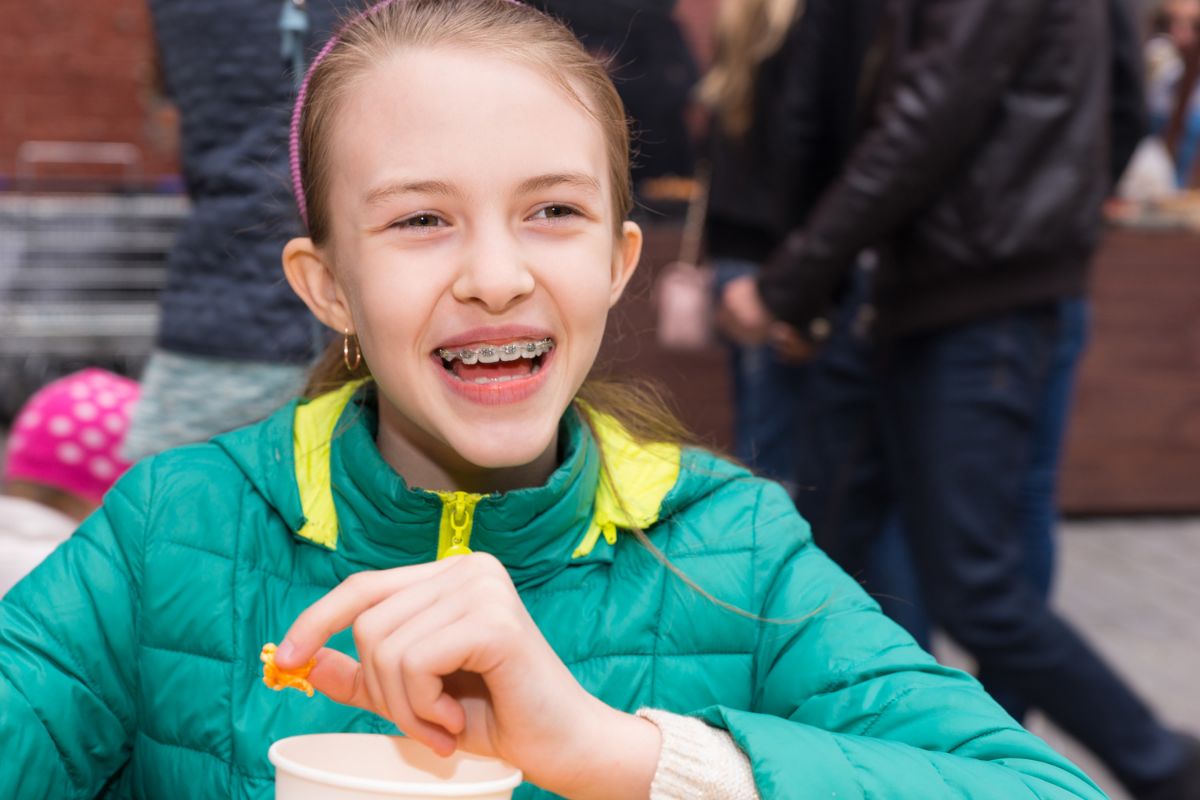 5 Stocking Stuffer Ideas for Kids with Braces Young child in green jacket smiling at camera while eating outdoors with people in background.