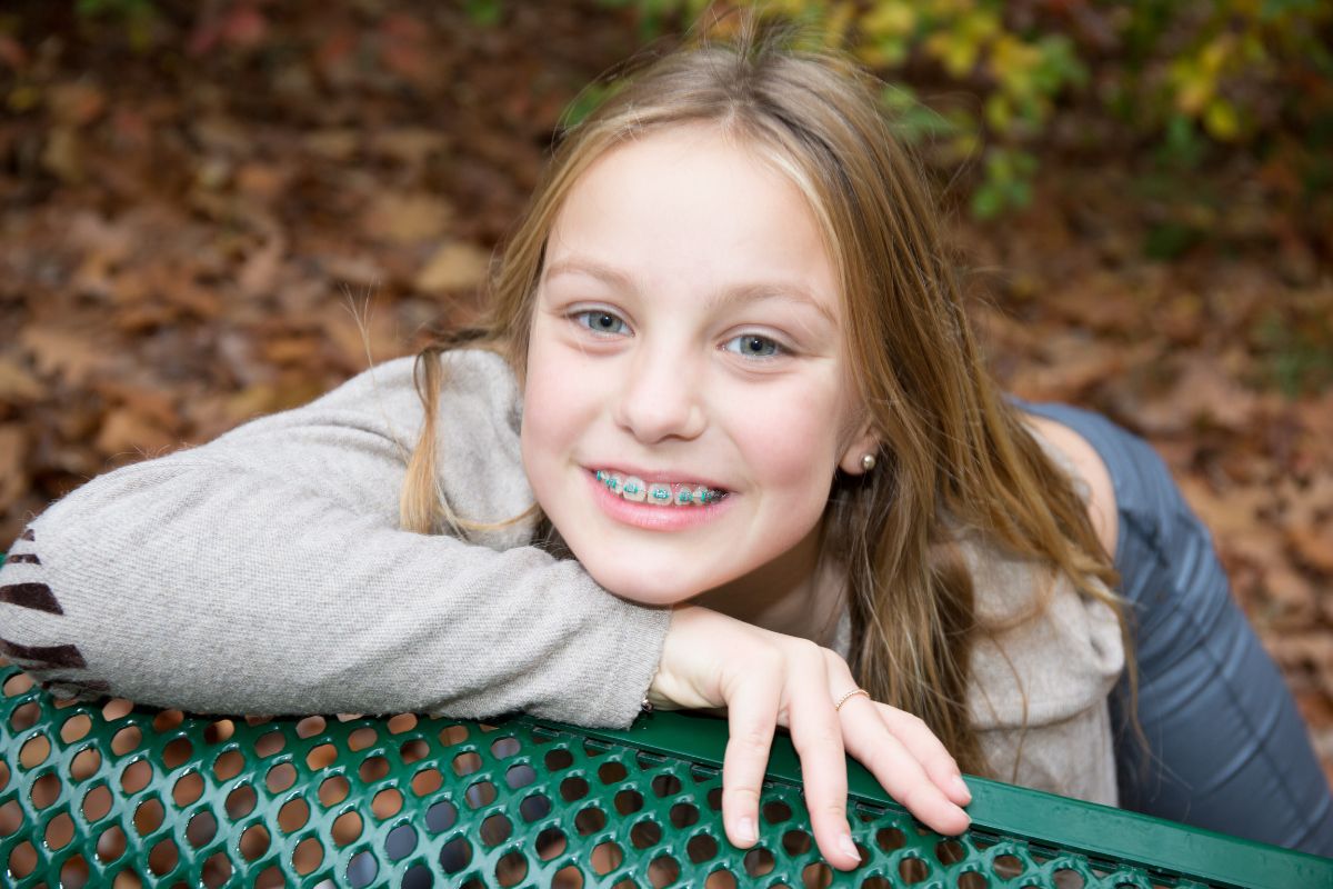 Young girl smiling at camera, leaning on green bench outdoors with autumn leaves background.